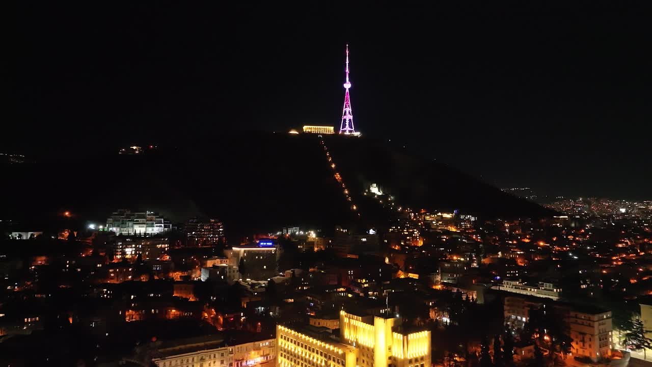 A magnificent communications tower illuminated in vibrant purple, standing tall over a glowing city at night. The scattered city lights form a mesmerizing pattern below, adding depth and beauty