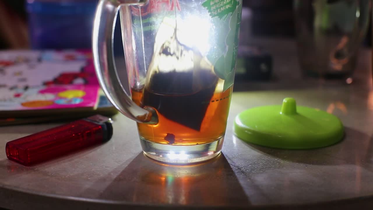 유리 뚜껑, 가스 라이터, 책 등의 배경에 대해 뜨거운 물이 가득 찬 유리 컵에 티백을 넣습니다.