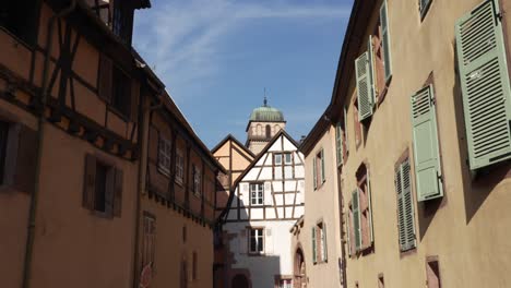 Edificios-Arquitectónicos-Con-Entramado-De-Madera-En-Kaysersberg,-Francia,-La-Torre-Del-Reloj-De-La-Iglesia-En-El-Fondo