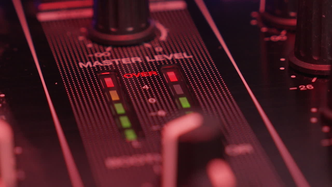 Nærbillede af DJ-mixerens hovedmålere, der rammer rødt og viser høj musik-audiooverbelastning i en mørk klub.