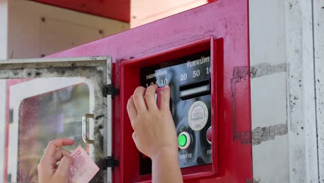 A person inserts cash into a red coin-operated fuel dispenser in Phuket, Thailand. Bright daylight illuminates the scene