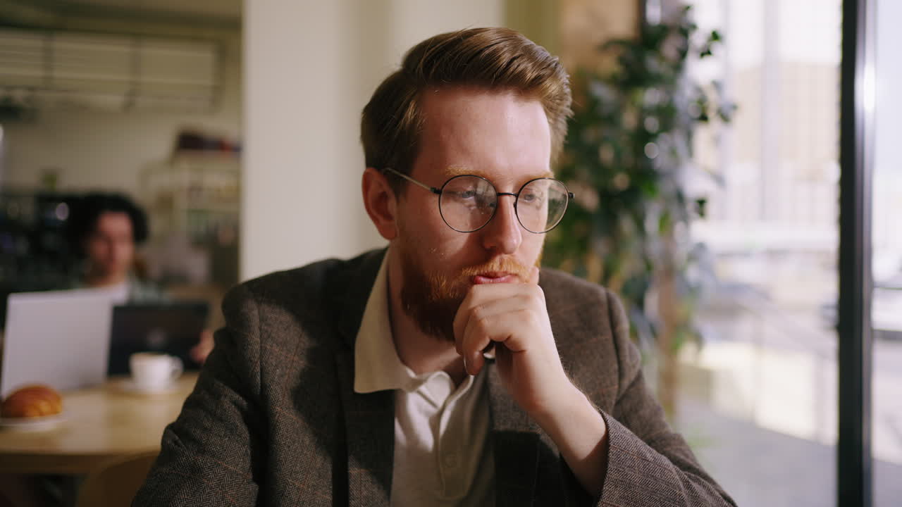 A man with a beard and glasses sits at a table in a cafe and works on his laptop.
