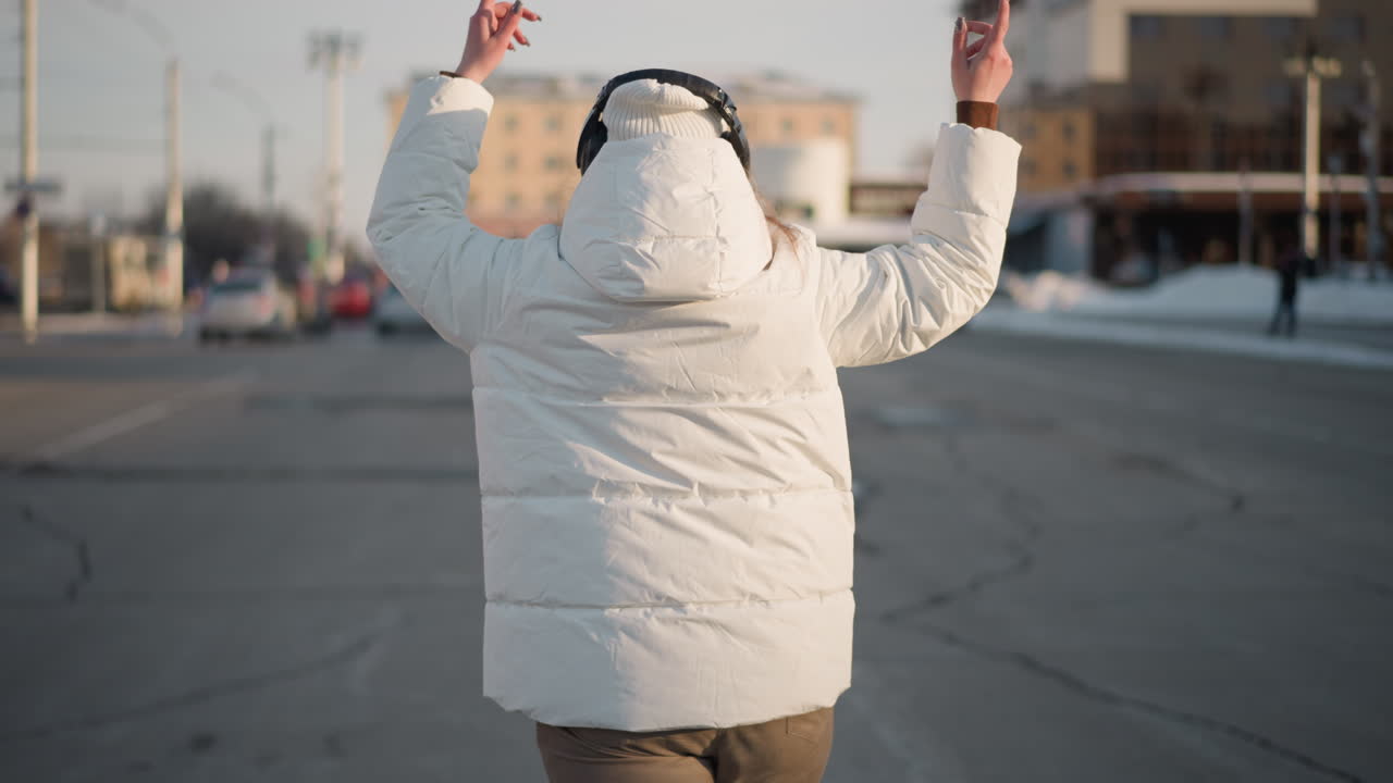 Back view of female in white coat swaying arms with headphones on ears, standing confidently in open street on cold winter day, immersed in music, radiating calm and freedom through motion