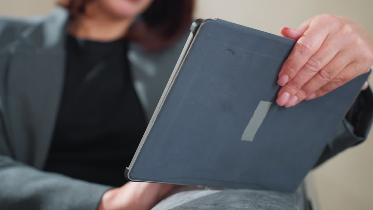 Businesswoman in grey suit holding digital tablet while gesturing with other hand, possibly during video call or touchscreen interaction, sitting indoors in calm environment with focus on productivity