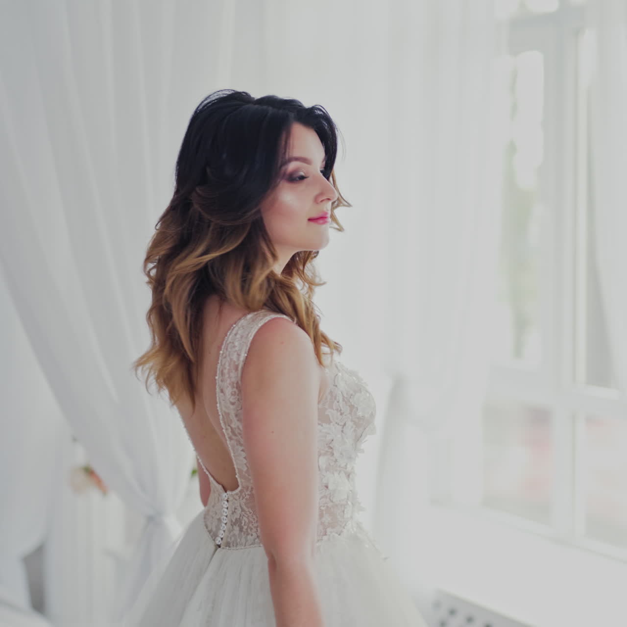 Sexy wedding lace dress on a beautiful model in the light studio. Attractive bride in luxurious dress with bouquet of flowers is coming to the window in bedroom.