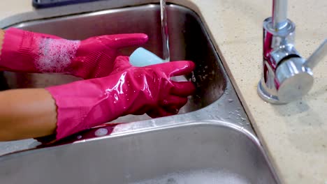 hands in pink gloves wash a cup under running water in a kitchen sink. bright lighting highlights the cleaning process