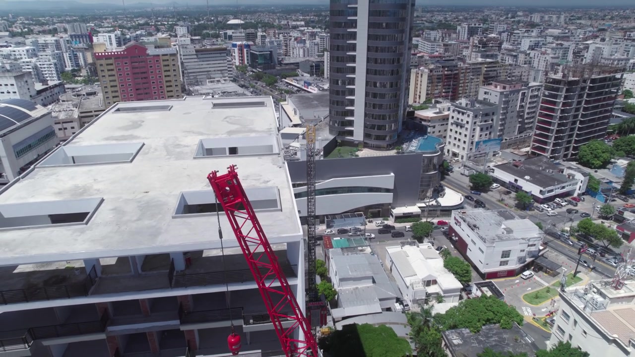 Tomada aérea de drone em ângulo baixo subindo e recuando ao lado de um guindaste telescópico e um edifício em construção, revelando a paisagem urbana comercial em Santo Domingo