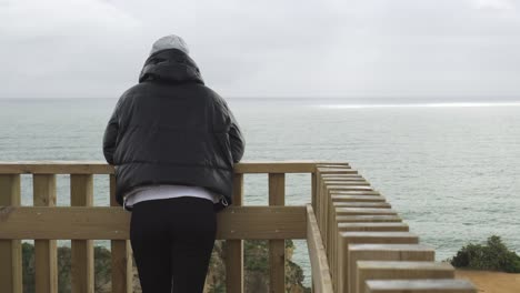chica con gorra y ropa oscura en un mirador de madera frente al mar mirando en silencio el paisaje marítimo