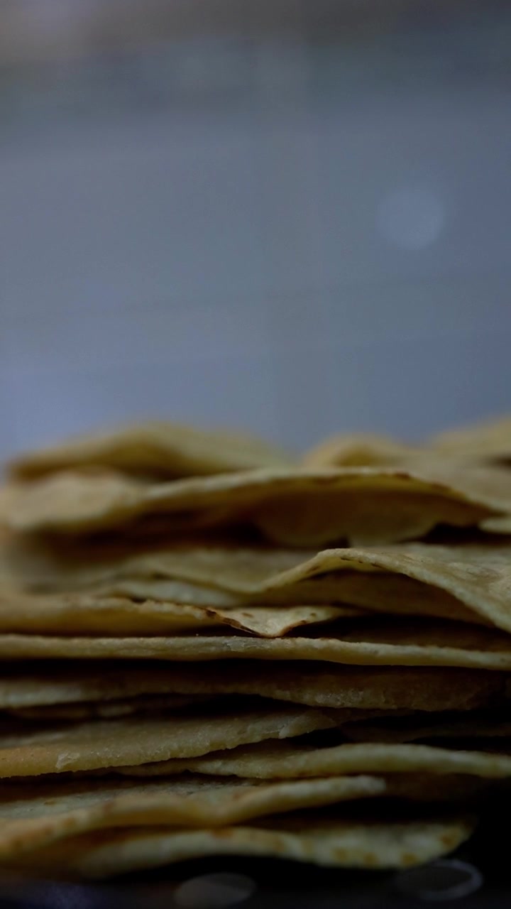 Stack of thin golden chapati flatbread in close-up with soft, blurred blue sky background. No people visible. Warm, earthy tones with shallow depth of field creating intimate, artistic mood.