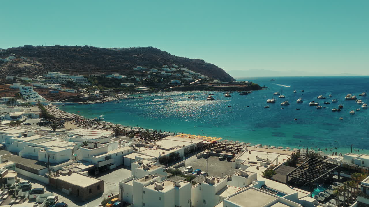 Drone panning above Mykonos showing white houses, winding streets, and kitesurfers on the blue Aegean Sea