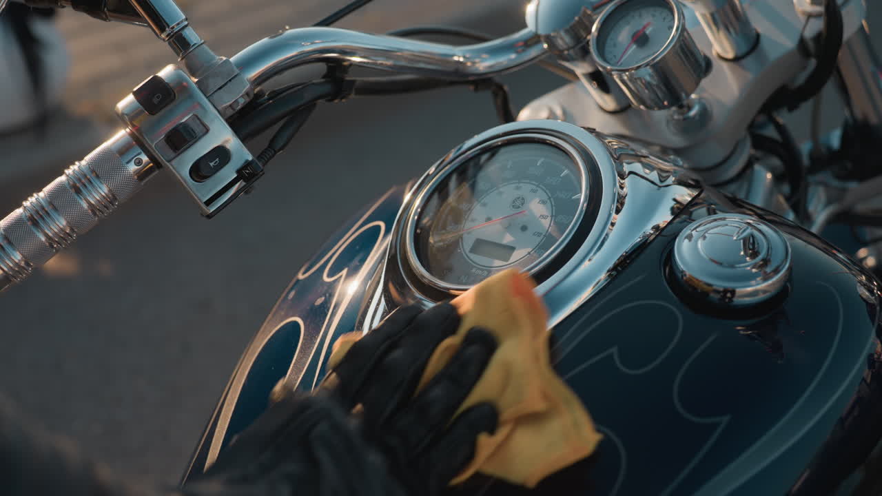 Biker in leather jacket uses yellow cloth to clean motorcycle speedometer and chrome tank, highlighting shiny details of handlebar and metallic parts under warm sunlight with clear reflections