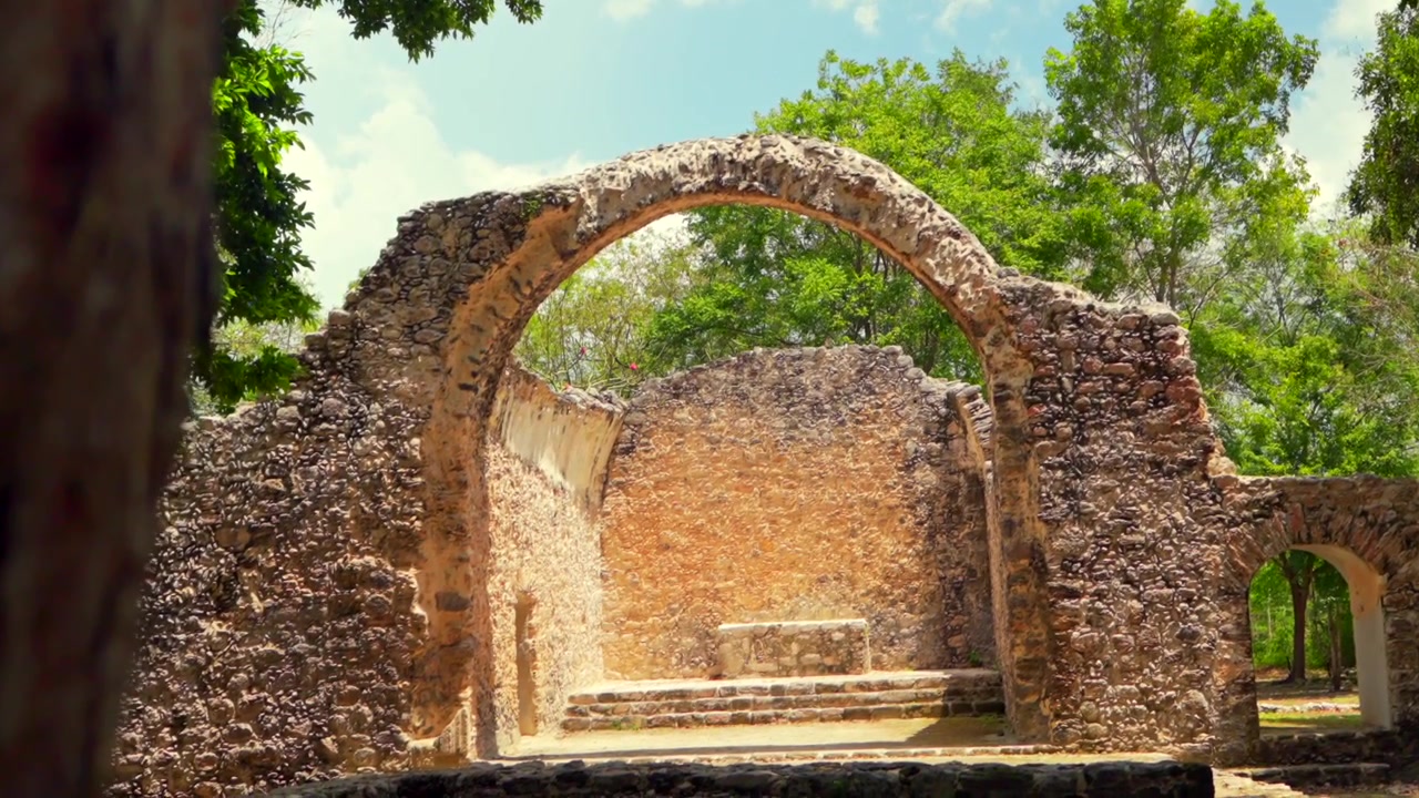 Una ripresa in avvicinamento accattivante rivela le storiche rovine della Cappella Spagnola presso il sito archeologico di Oxtankah. Questa antica struttura costruita con pietre Maya è circondata da alberi verdi lussureggianti sotto un cielo blu.
