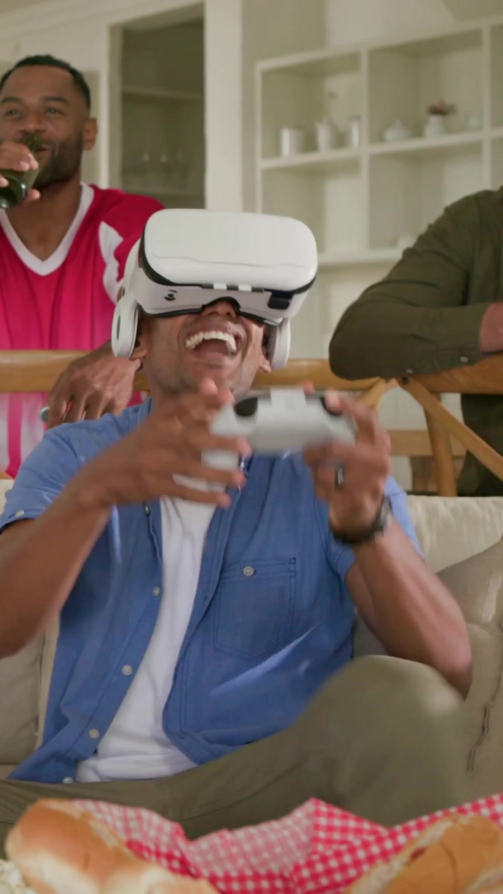 Video verticale: Uomo in maglietta blu con visore VR che inizia una partita giocando con un controller a casa, amici con birra