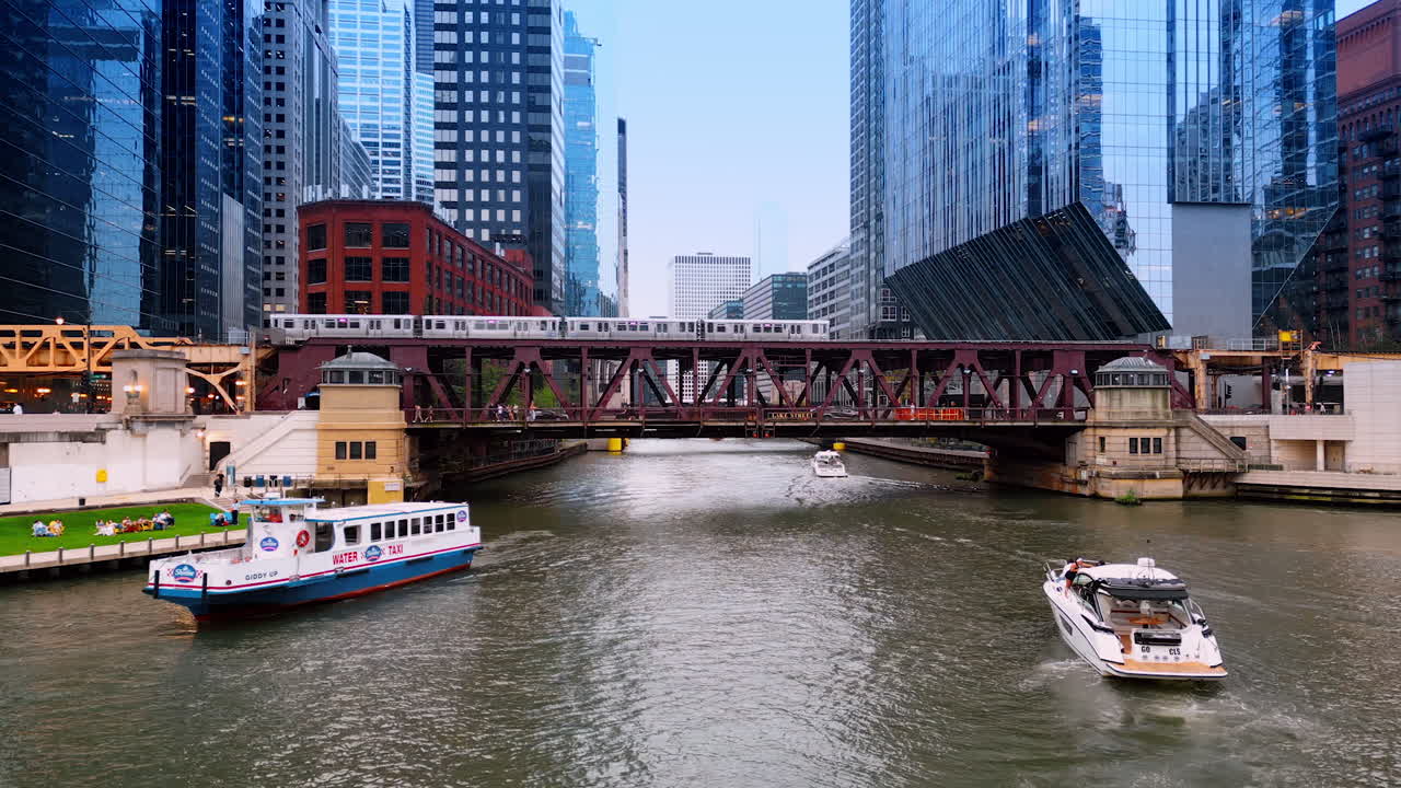 Chicago, USA, 29 June 2025: Three boats move by the waterscape of the Chicago River. A train goes by the bridge over the river. Low angle view at the glass skyscrapers on the waterfronts