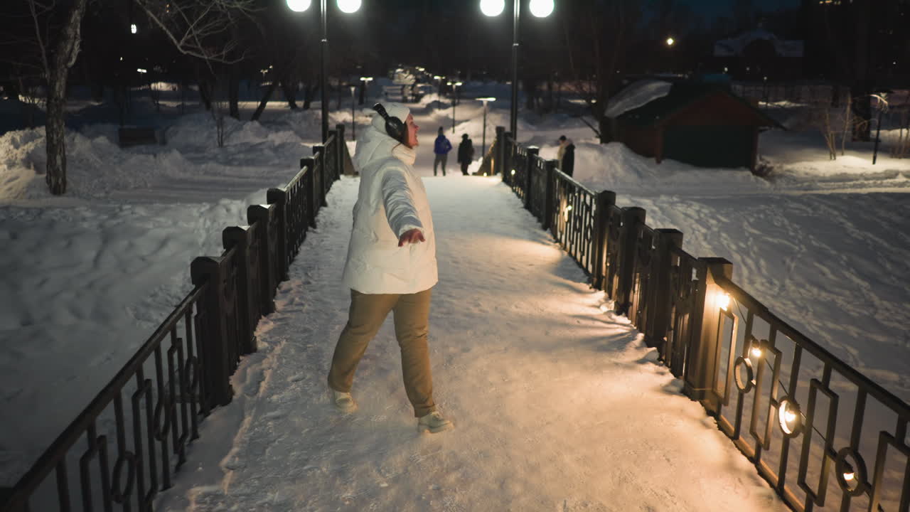 Young artist wearing white puffer coat and headphones dances passionately on snow covered walkway illuminated by lampposts and string lights in park while passersby stroll under winter night sky