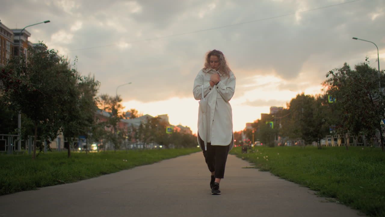 Cold girl covering herself strolling on paved path with shimmering car headlamps in background urban setting during sunset with cloudy sky greenery buildings roadside signposts and soft evening light
