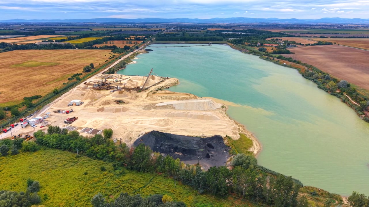 Aerial perspective of sand mining pit and turquoise reservoir. Dark coal or sediment deposits sit alongside sand piles on the bank of an expansive green lake.