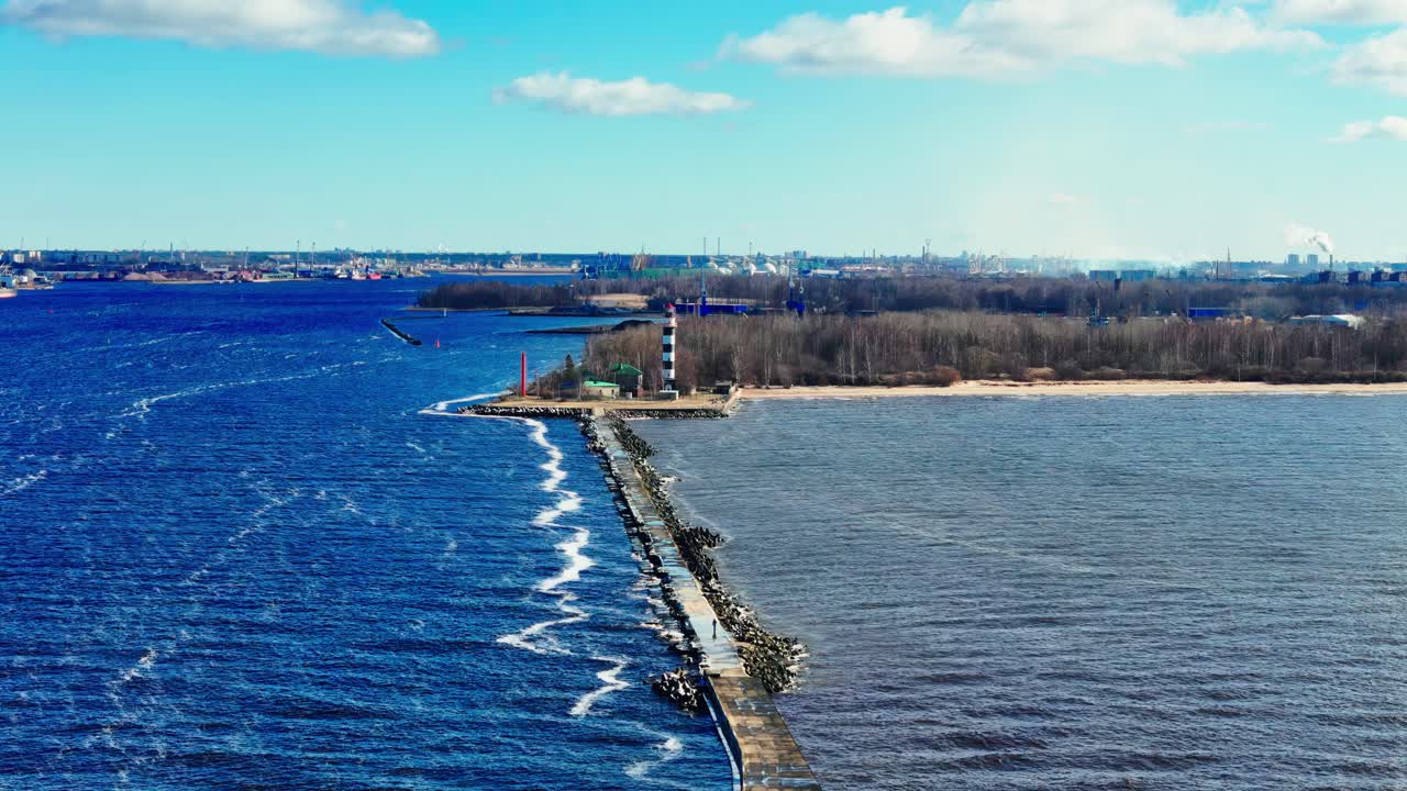 Aerial view of a slender stone jetty separating choppy blue sea from tranquil estuary waters, leading to a lighthouse and forested coastline under a bright sky.