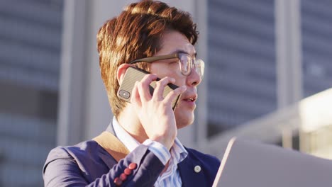 Asiatischer-Mann-Spricht-Auf-Dem-Smartphone,-Während-Er-Im-Firmenpark-Einen-Laptop-Benutzt