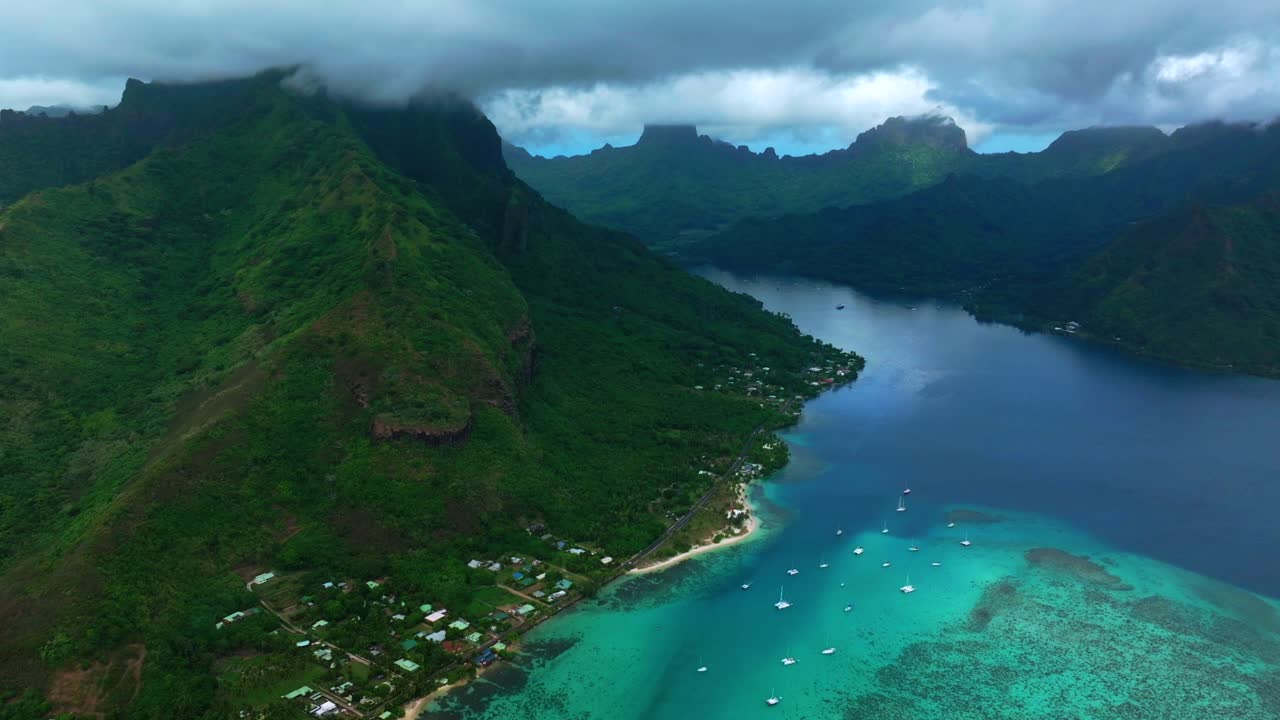 Moorea, Polynesia thuộc Pháp, quang cảnh từ drone trên không với bờ biển rải bungalow, Vịnh Opunohu và làn nước đầm phá màu ngọc lam sáng bên dưới những sườn đảo xanh tươi cùng những chiếc thuyền buồm neo đậu di chuyển vòng tròn sang phải
