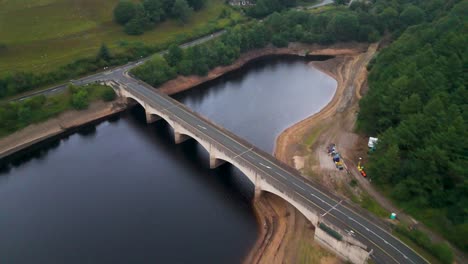 drone footage glides above a multi-arched bridge spanning calm reservoir waters, surrounded by lush greenery and overcast natural lighting in hope valley, england