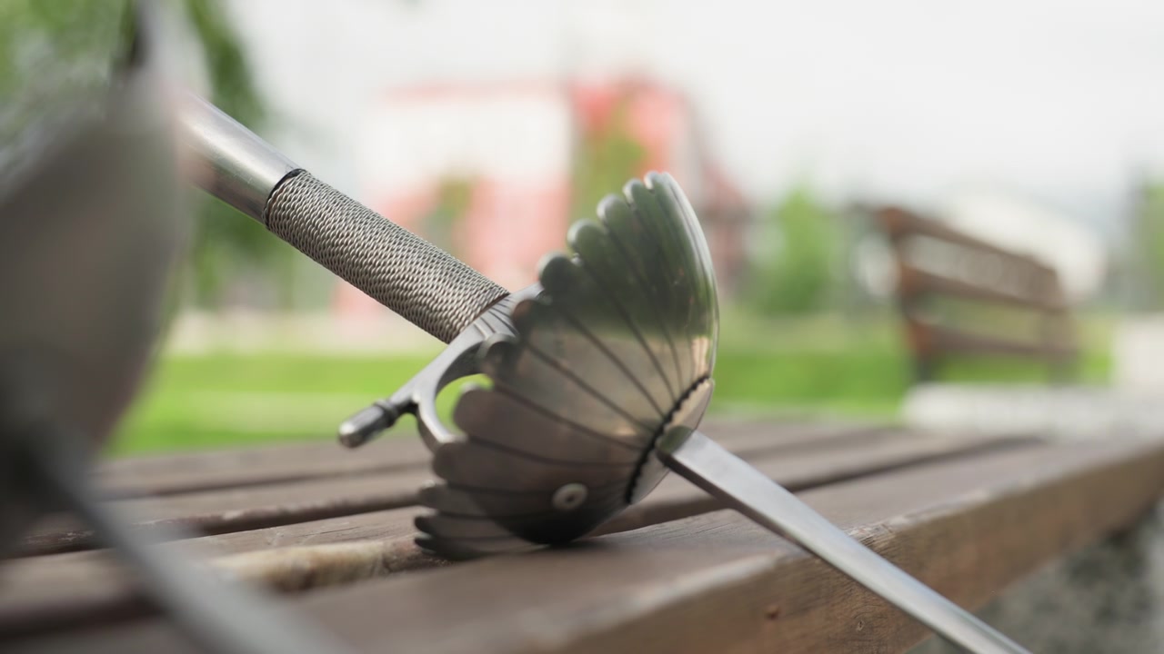 Nærbillede af en hånd, der placerer og justerer to fægtesværd på trælameller i et udendørs parkmiljø. Metalbladene hviler stabilt og fremhæver deres detaljerede konstruktion mod en sløret baggrund.