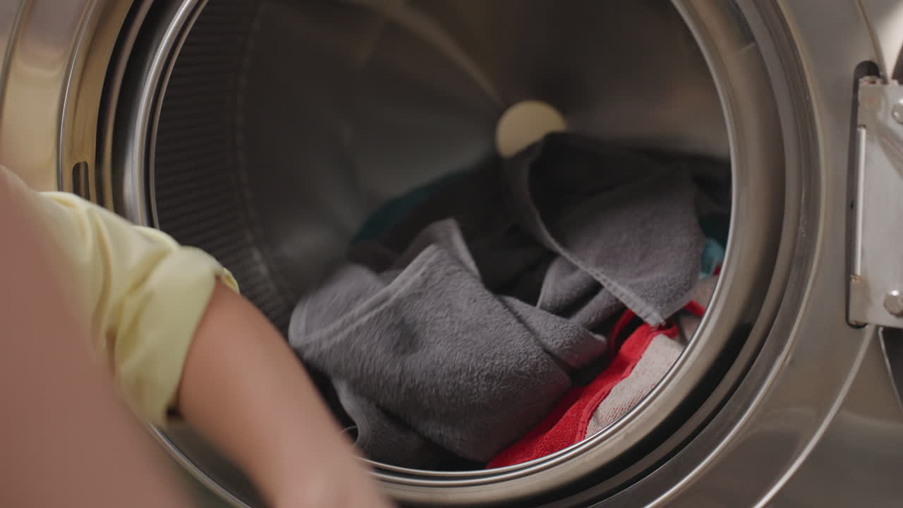 Close view inside washer shows laundry woman sorting pile, placing garments into drum with steady hands, gray towel foreground, colorful fabrics visible, stainless interior