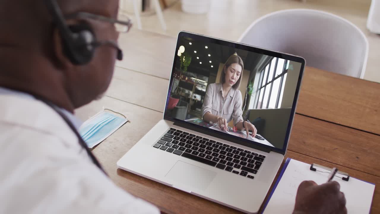 Afroamerikansk mandlig læge med headset på, der tager noter under et videoopkald på sin bærbare computer