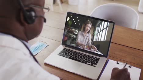 Afroamerikanischer-Männlicher-Arzt-Mit-Telefon-Headset,-Der-Notizen-Macht,-Während-Er-Einen-Videoanruf-Auf-Dem-Laptop-Führt