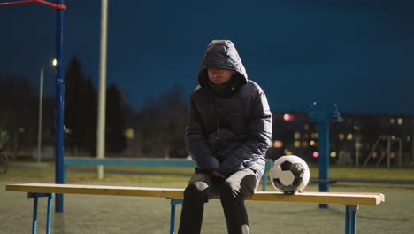 a thoughtful man sits alone on a bench outdoors, with his hands clasped between his legs, a soccer ball rests beside him, with someone riding a bicycle in the background