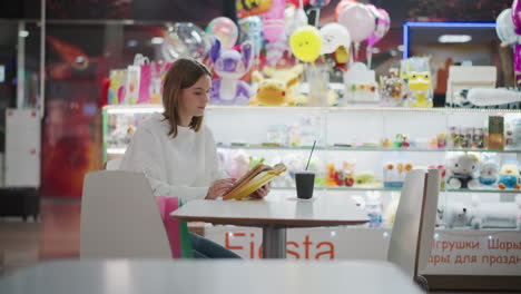 lady sitting in mall reading yellow book, with coffee cup and colorful shopping bags on table, soft lighting surrounds her, with vibrant toy displays and blurred background