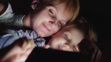 Happy-Mother-With-Her-Daughter-Together-I-Play-On-The-Tablet-Lie-Side-By-Side-Face-To-Face-In-Bed