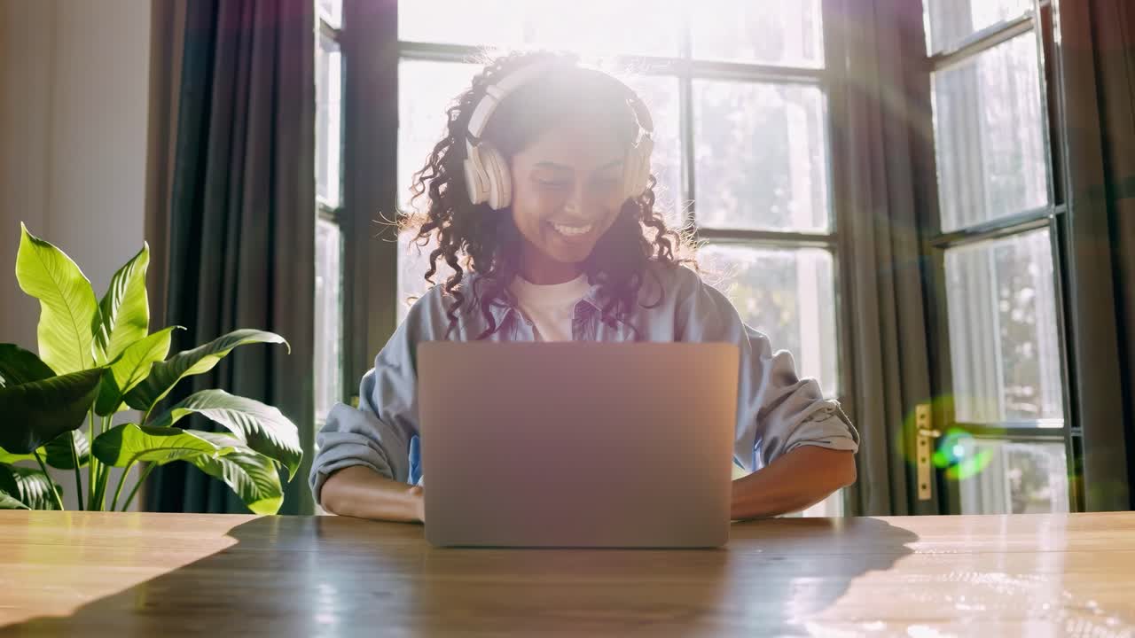 A woman wearing headphones smiles at her laptop in a sunlit room. The low-angle shot adds a dynamic