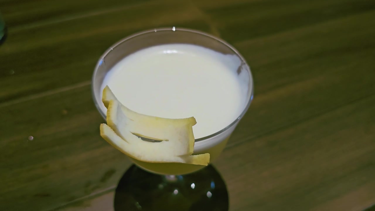 Primo piano di un cocktail giallo pallido in un bicchiere coupe con schiuma cremosa e guarnizione di scorza di agrumi, poggiato su un bancone di legno sotto una calda illuminazione da lounge per video di bevande artigianali