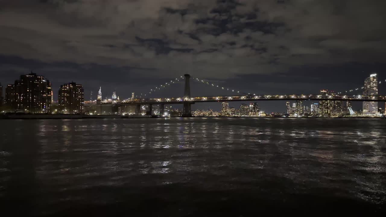 A tranquil nighttime view of the East River with the Manhattan Bridge illuminated against the backdrop of the city's skyline, capturing the serene beauty of the urban night