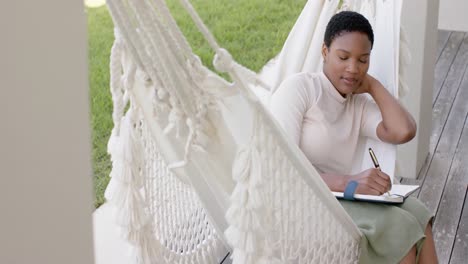 Glückliche-Afroamerikanische-Frau-Sitzt-In-Der-Hängematte-Auf-Der-Terrasse,-Schreibt-In-Notizbuch,-Zeitlupe