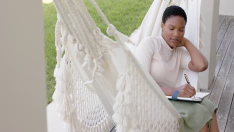 Feliz-Mujer-Afroamericana-Sentada-En-Una-Hamaca-En-La-Terraza,-Escribiendo-En-Un-Cuaderno,-Cámara-Lenta