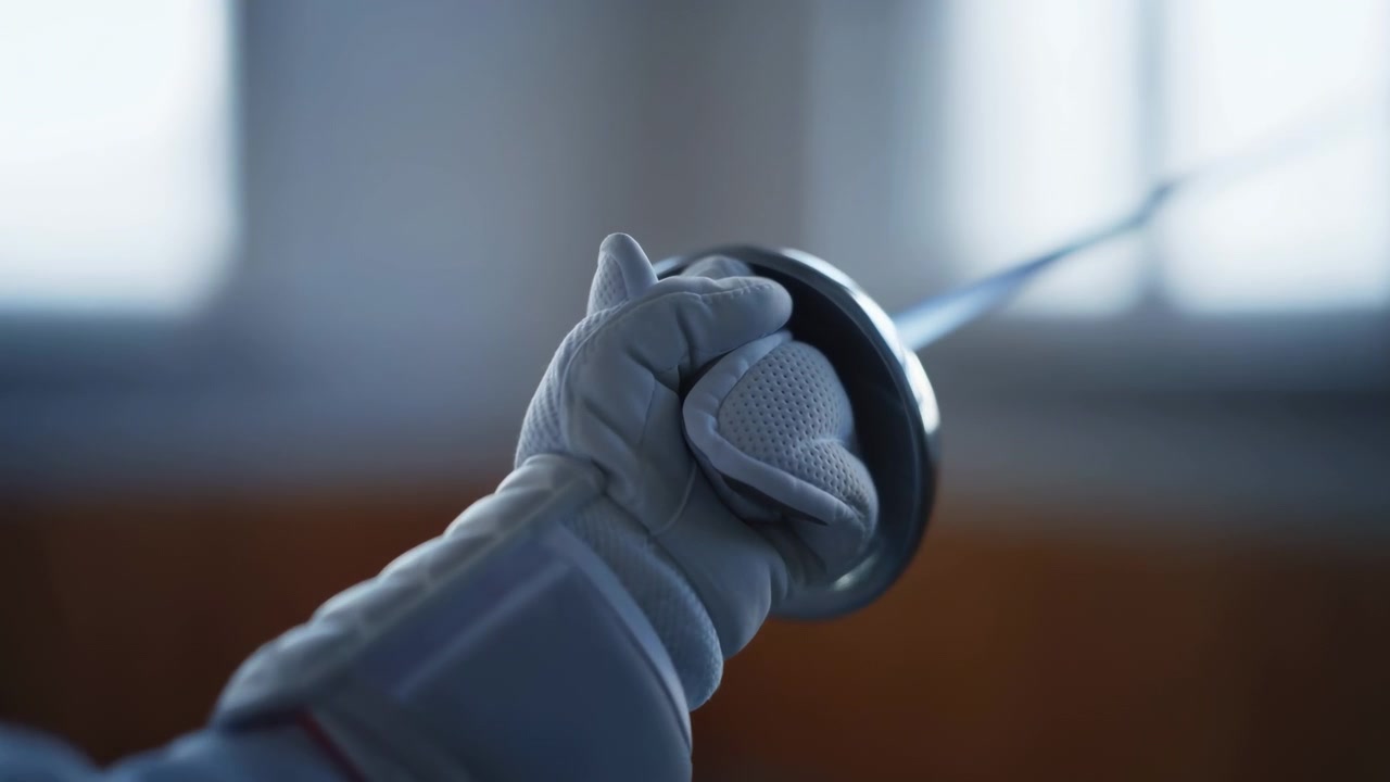 Close over shoulder view of fencer gripping foil hilt inside circular guard, blade extended toward right in indoor salle, with gloved hand, white jacket, and soft window light, with fencer, foil