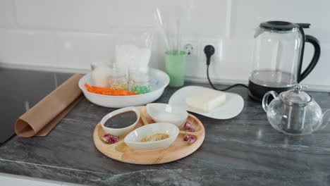 modern kitchen countertop with ceramic bowls containing spices, sugar, and sauce on wooden tray, butter on plate, glass jars with ingredients, glass kettle, and teapot