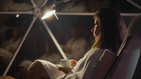 woman sits in armchair with cup of hot beverage in room lit by light of lantern. sad lady rests alone thinking about life problems. concept of depressed emotions