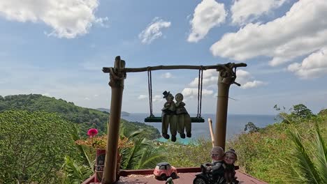 a swing with statues overlooks lush greenery and ocean views in phuket, thailand, under bright daylight