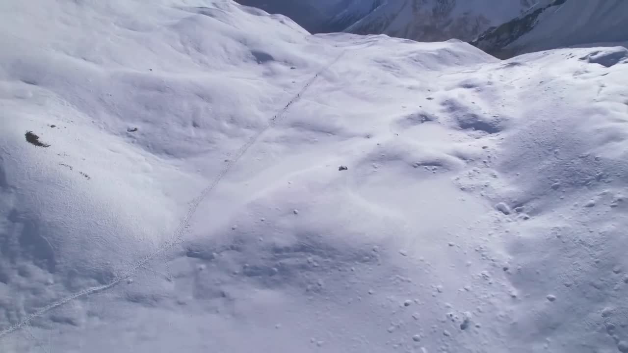 Aerial drone footage flying closer to white snow covered ground on top of a mountains and closer to a hiking track where hikers are walking on in Tibetan plateau. Footprints on the snow at mountains.
