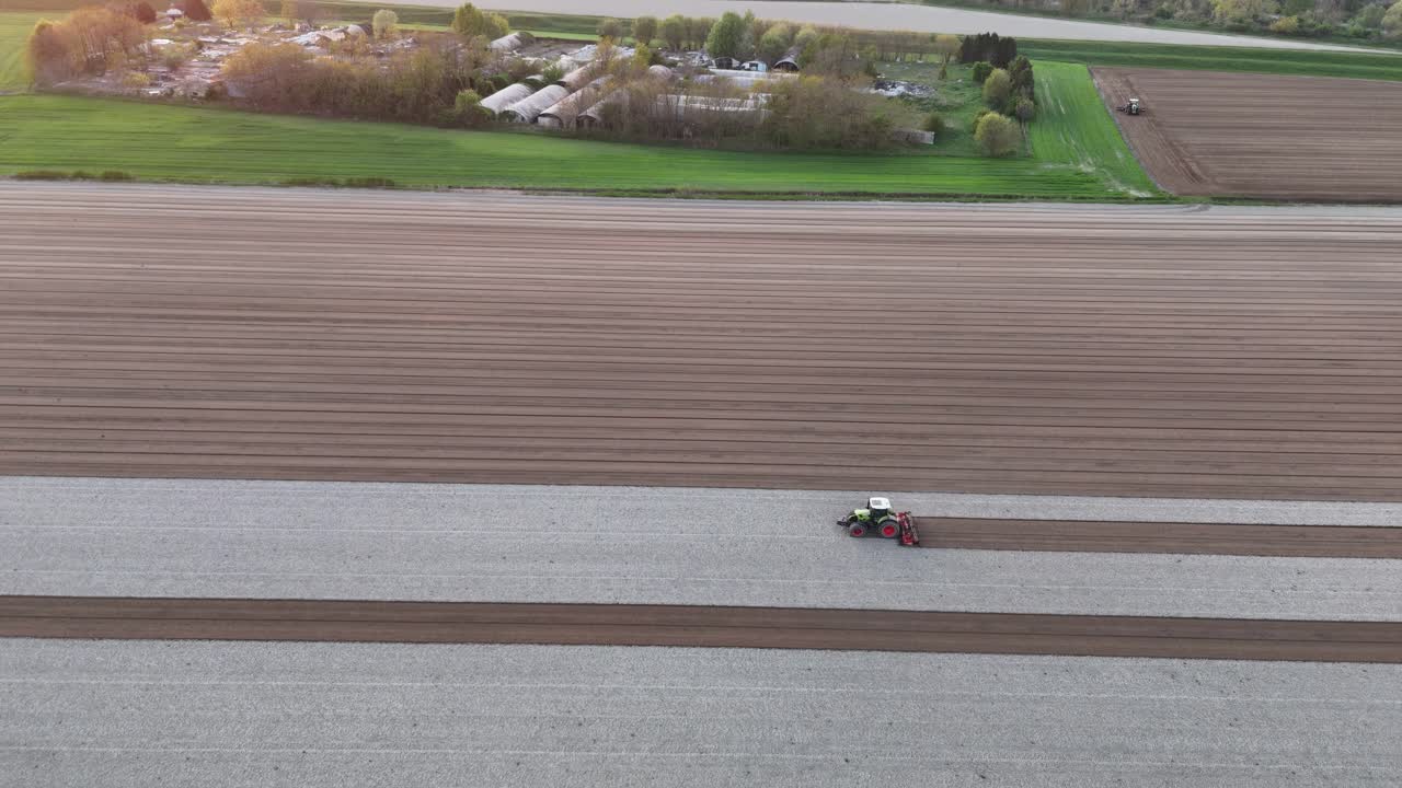 slow paning drone captures tractor pulling leveling harrow as it carves deep brown seed rows through bleached soil, set against the green edge of a farmyard and distant fields at golden hout light