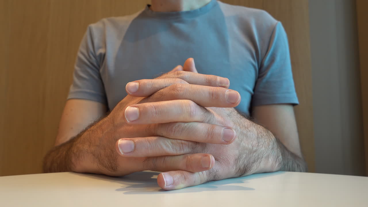 Man in grey shirt clasping his hands impatiently on a table