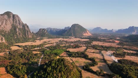 scenic drone glide above vang vieng, laos, showing limestone karst peaks, dry-season rice fields, scattered farms, and rural roads under warm golden light across the nam song valley