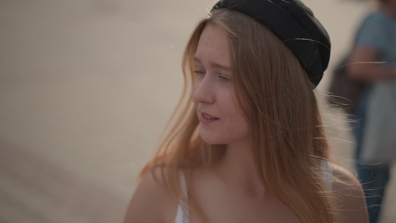 giovane donna caucasica con cuffie in una strada di città illuminata dal sole, sorriso spensierato e capelli fluenti, berretto e abbigliamento estivo, folla del festival sfocata sullo sfondo, bokeh dorato delicato, ritratto spontaneo