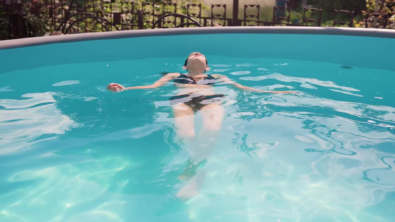 Woman floating on her back in clear blue pool with sunlight reflections showing relaxed posture and calm water ripples. Pool edge visible above woman head background includes greenery