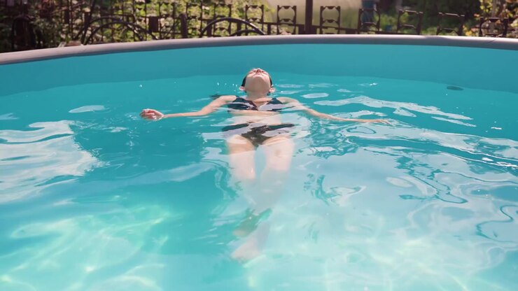 Woman floating on her back in clear blue pool with sunlight reflections showing relaxed posture and calm water ripples. Pool edge visible above woman head background includes greenery