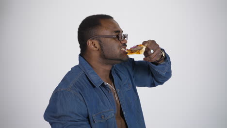 black man in glasses eats pizza. afro american man in blue shirt tastes a piece of delicious pizza on white studio background. profile view.