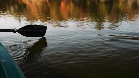 a close-up of a kayak paddle cutting through calm water, creating ripples on the lake’s surface as autumn colors reflect in the background